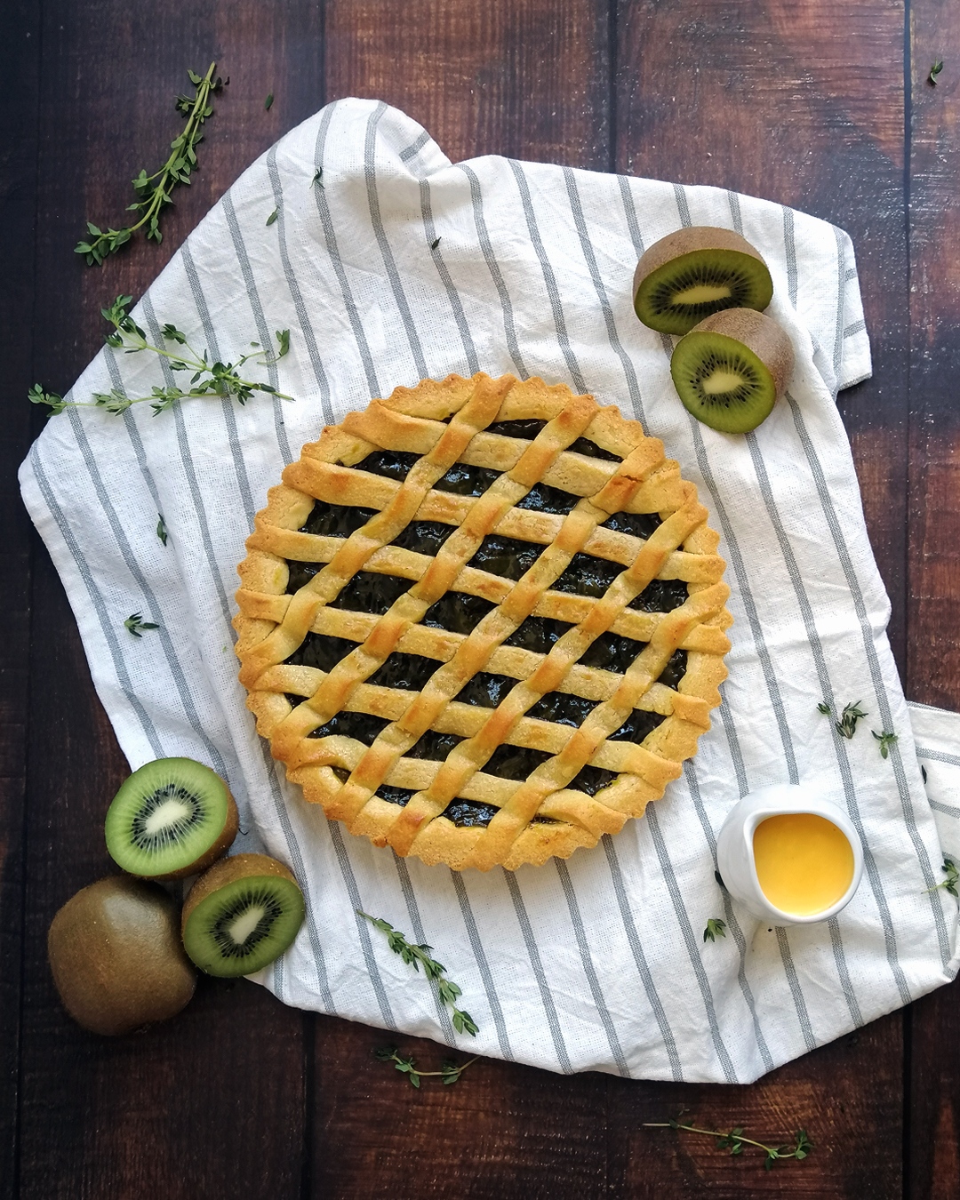 Kiwi compote baked tart with criss cross pastry top, laying on a striped tea towel. Fresh kiwis and jug of custard laying around