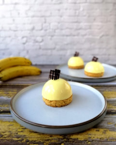 Banana and vanilla shiny light yellow dome sitting on a biscuit base , topped with a dark chocolate square decoration