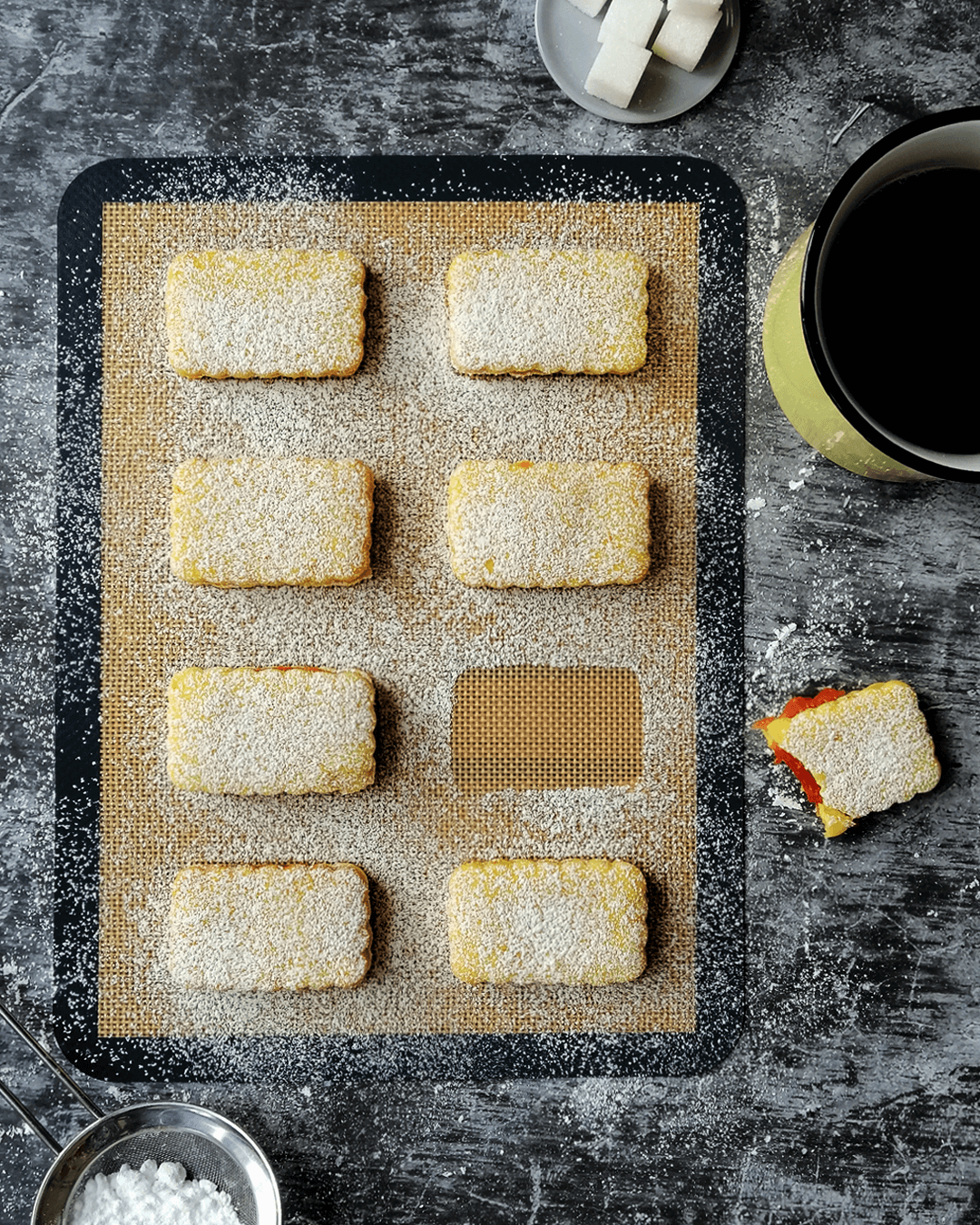 Rhubarb-filled orange biscuits lined on a baking mat, surrounded by a cup of tea, half eaten biscuit