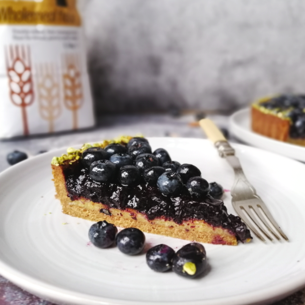 A juicy slice of blueberry and almond frangipane tart, laying on a plate and showing the wholewheat whortcrust pastry, almond frangipane, blueberry compote and fresh blueberries