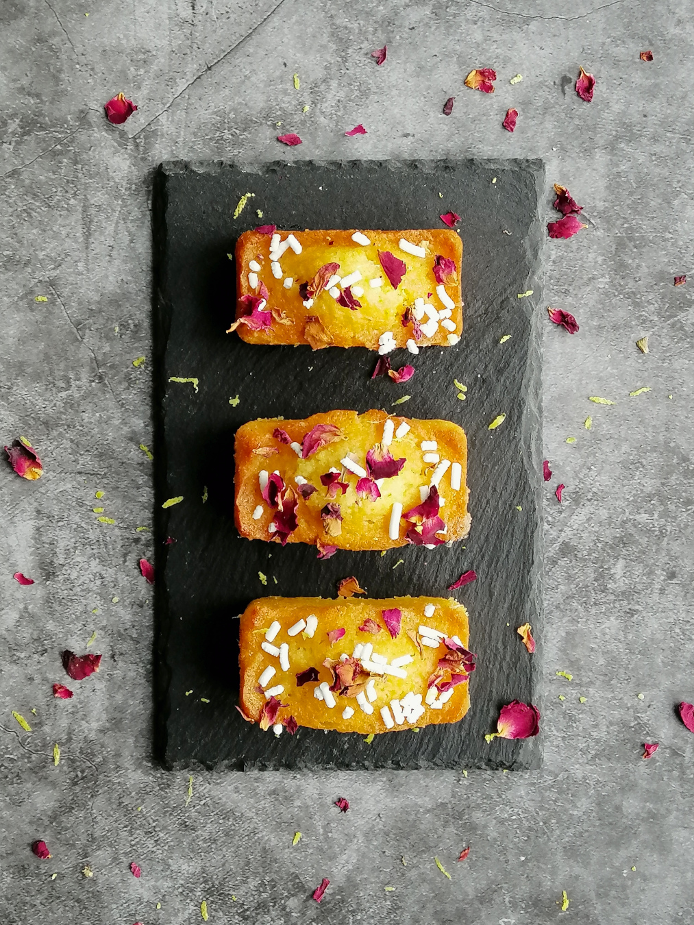 three lemon and rose mini drizzle cakes seen from the top, lined up on a grey slate